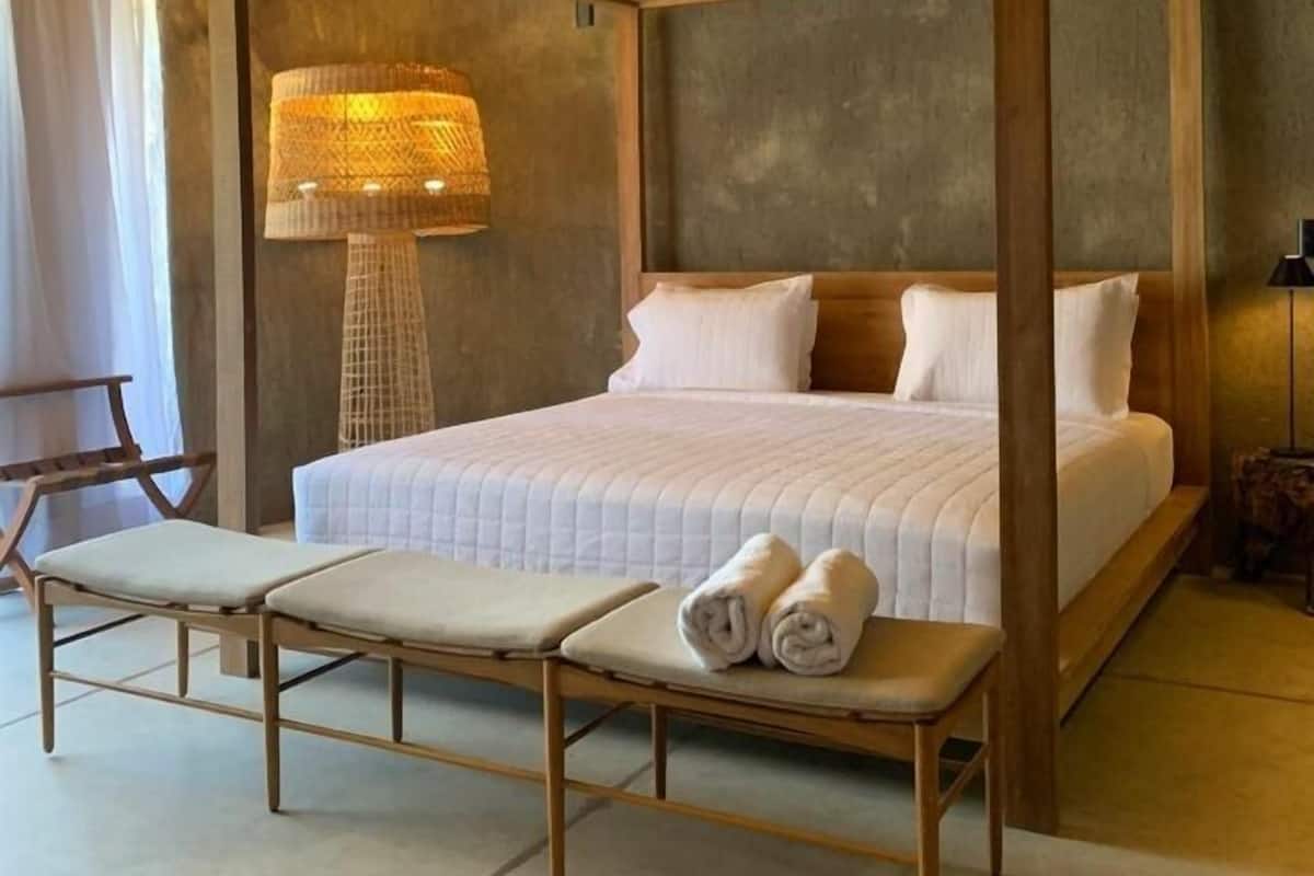 A four-poster bed with a white bedspread, two rolled towels on a bench, and a woven lamp beside the bed.