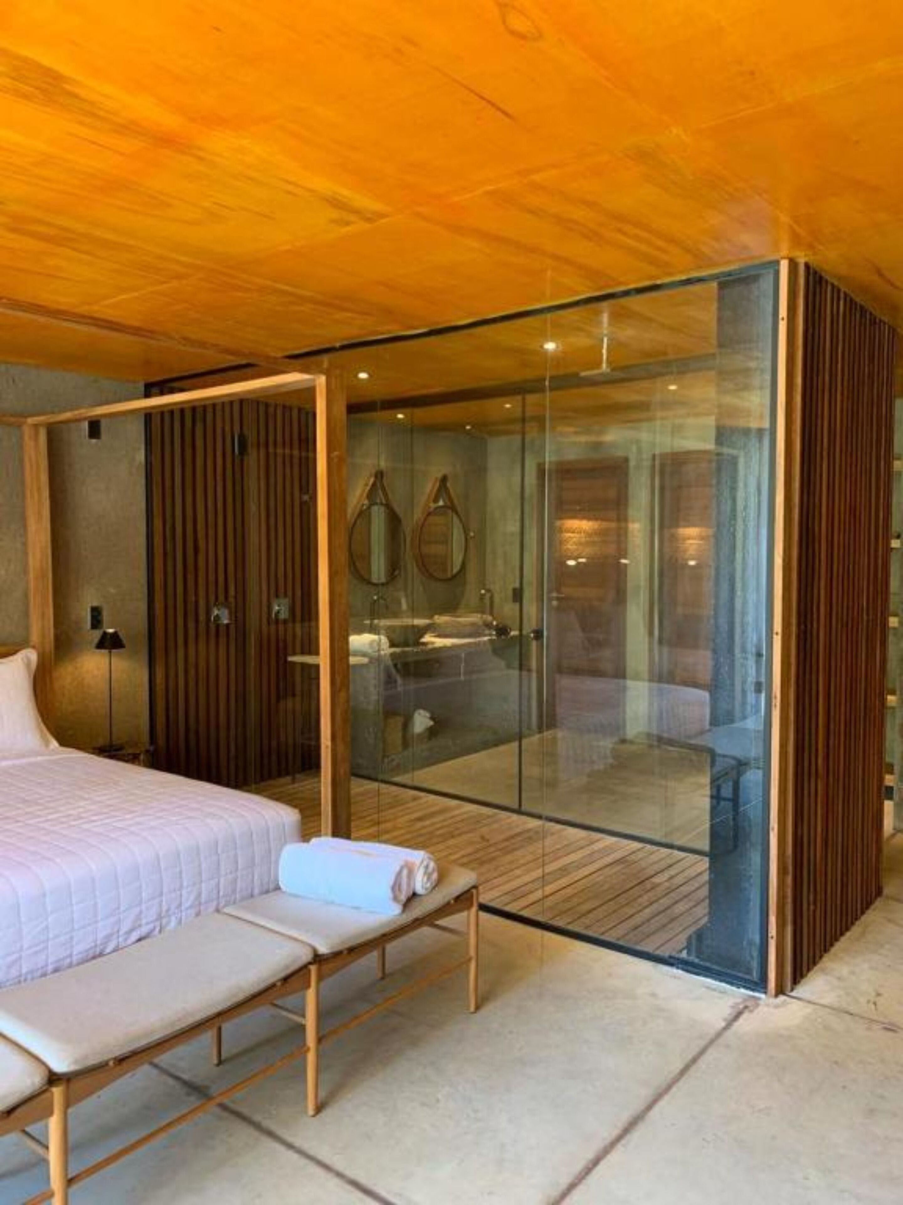 Ein modernes Schlafzimmer mit Glastrennwand, einem Bett, einer Bank und einem sichtbaren Badezimmer mit Waschbecken und Spiegel.