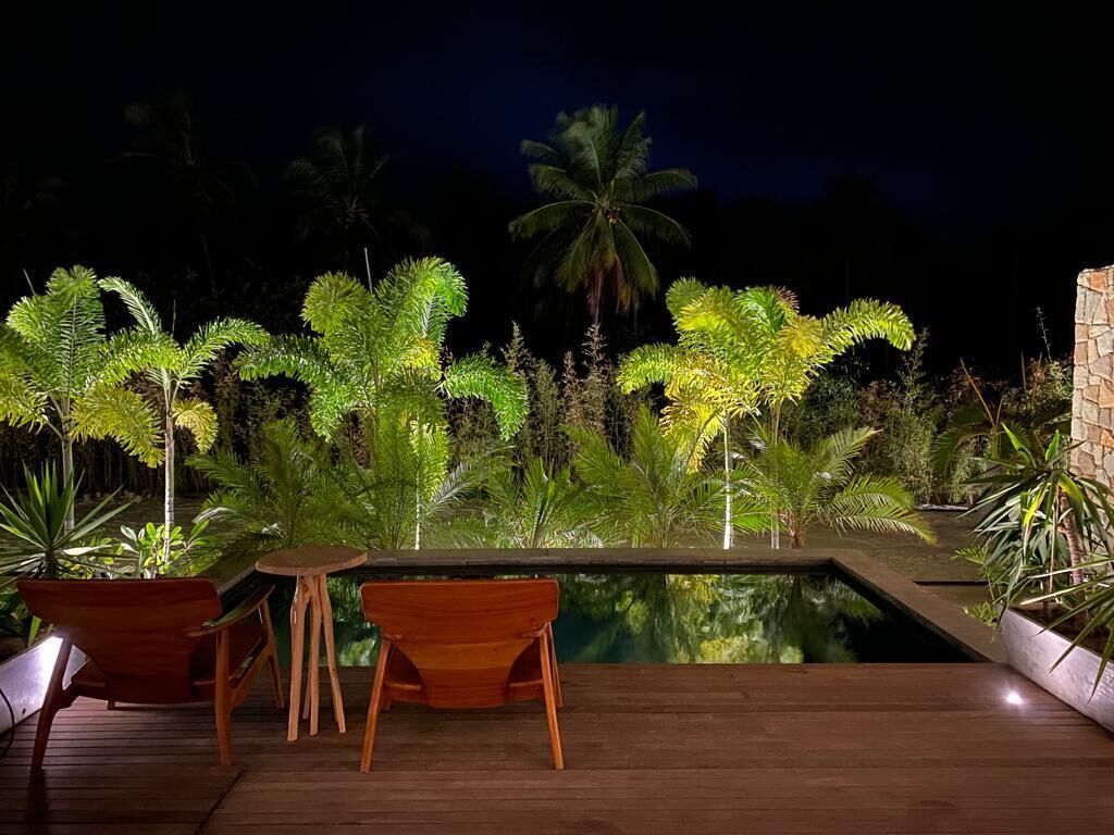 A nighttime outdoor area with a pool, wooden furniture, and tropical plants.