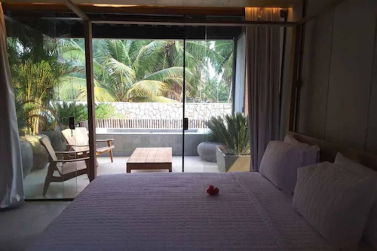 A bedroom with a bed, a small table, and a chair. There is a balcony with a view of palm trees.