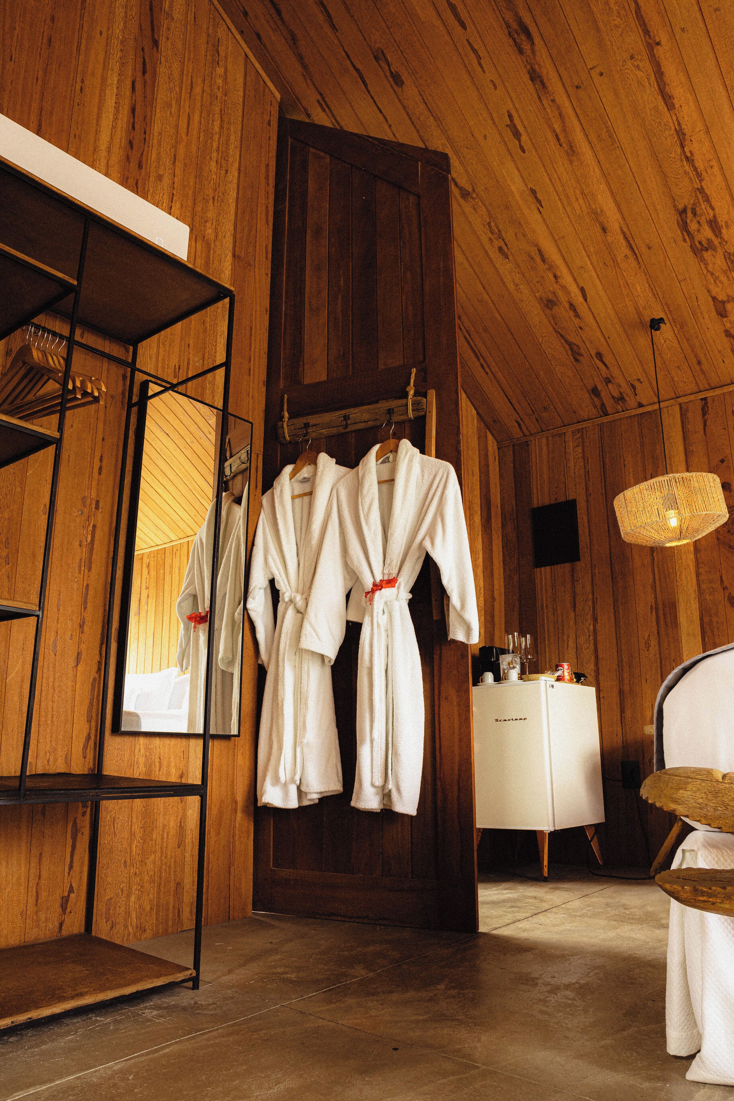 A wooden interior with a mirror, two white robes hanging, a small white table, and a wicker lamp.