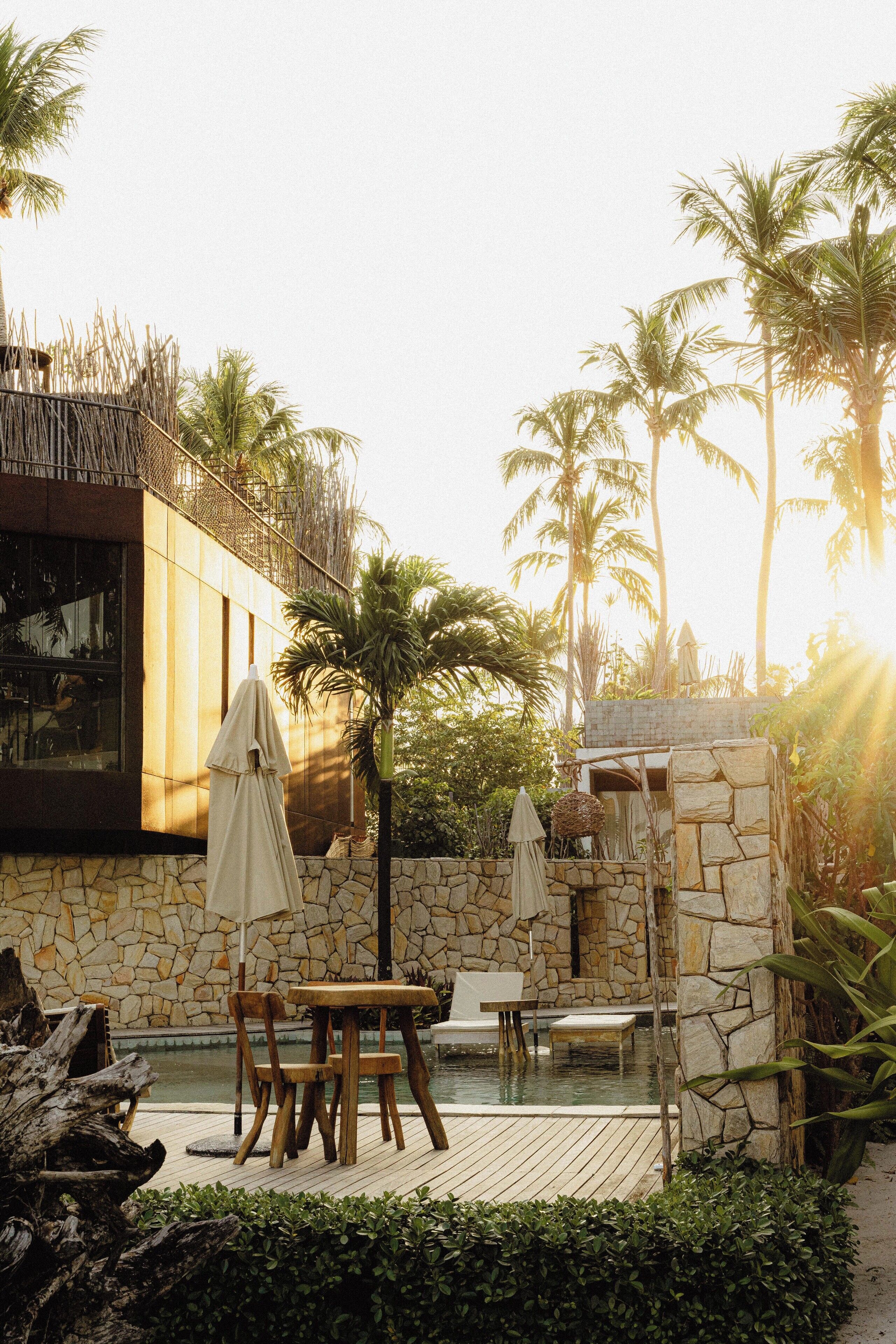 An outdoor patio area with stone walls, wooden furniture, and palm trees.