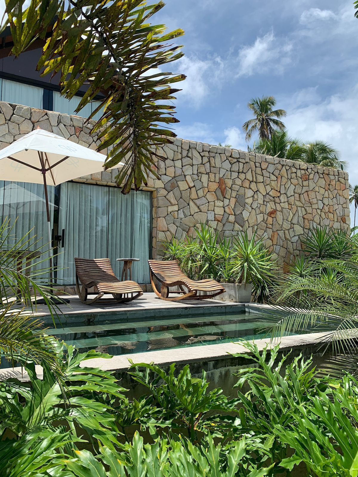 Un área de piscina con sillones de descanso y una sombrilla, rodeada de plantas tropicales y un muro de piedra.