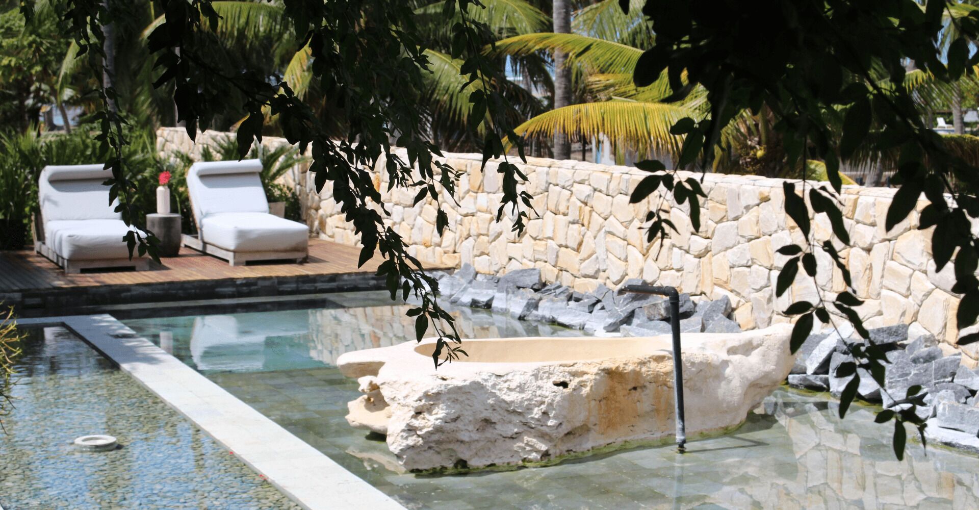 A pool with a stone wall and a waterfall feature, surrounded by lounge chairs and tropical plants.