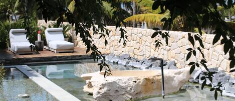 Una piscina con pared de piedra y una cascada, rodeada de sillas de descanso y plantas tropicales.