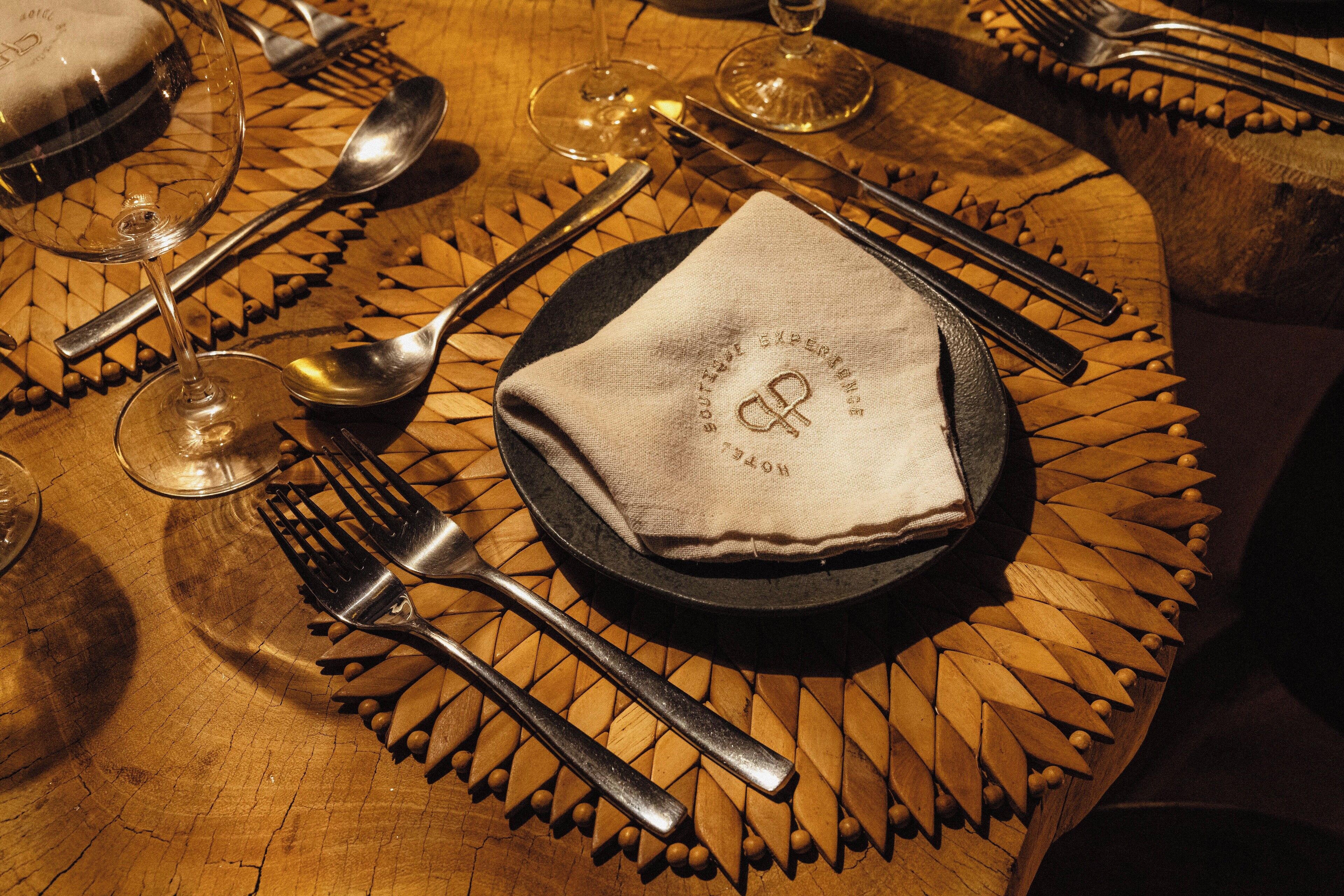 Une table ronde avec un set de table en fibres tressées, une serviette pliée ornée d’un logo et des couverts disposés pour le repas.