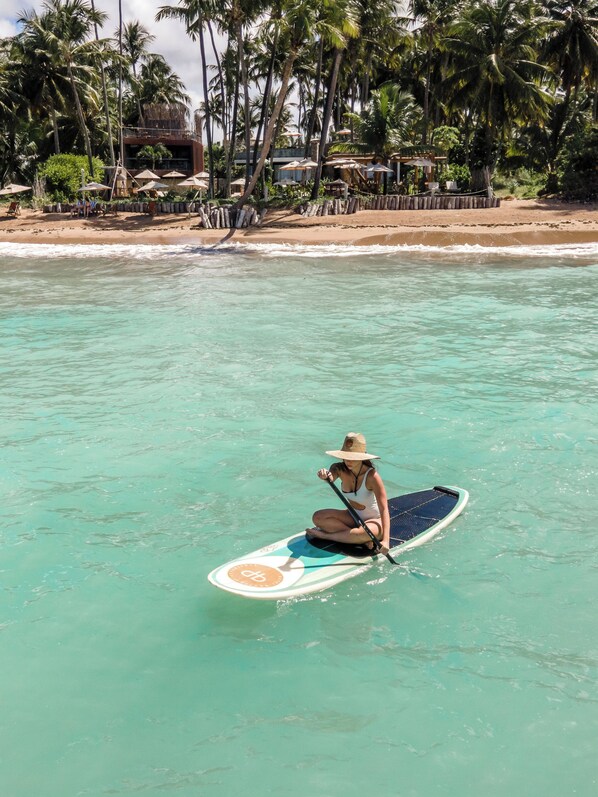 Una persona haciendo paddleboard sobre aguas turquesas cristalinas, con una playa y palmeras al fondo.