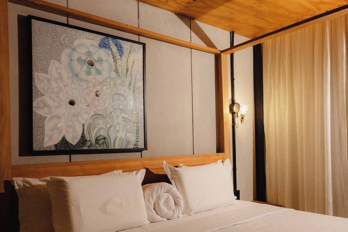 A neatly made bed with white linens, a wooden headboard, and a large floral artwork on the wall above it.