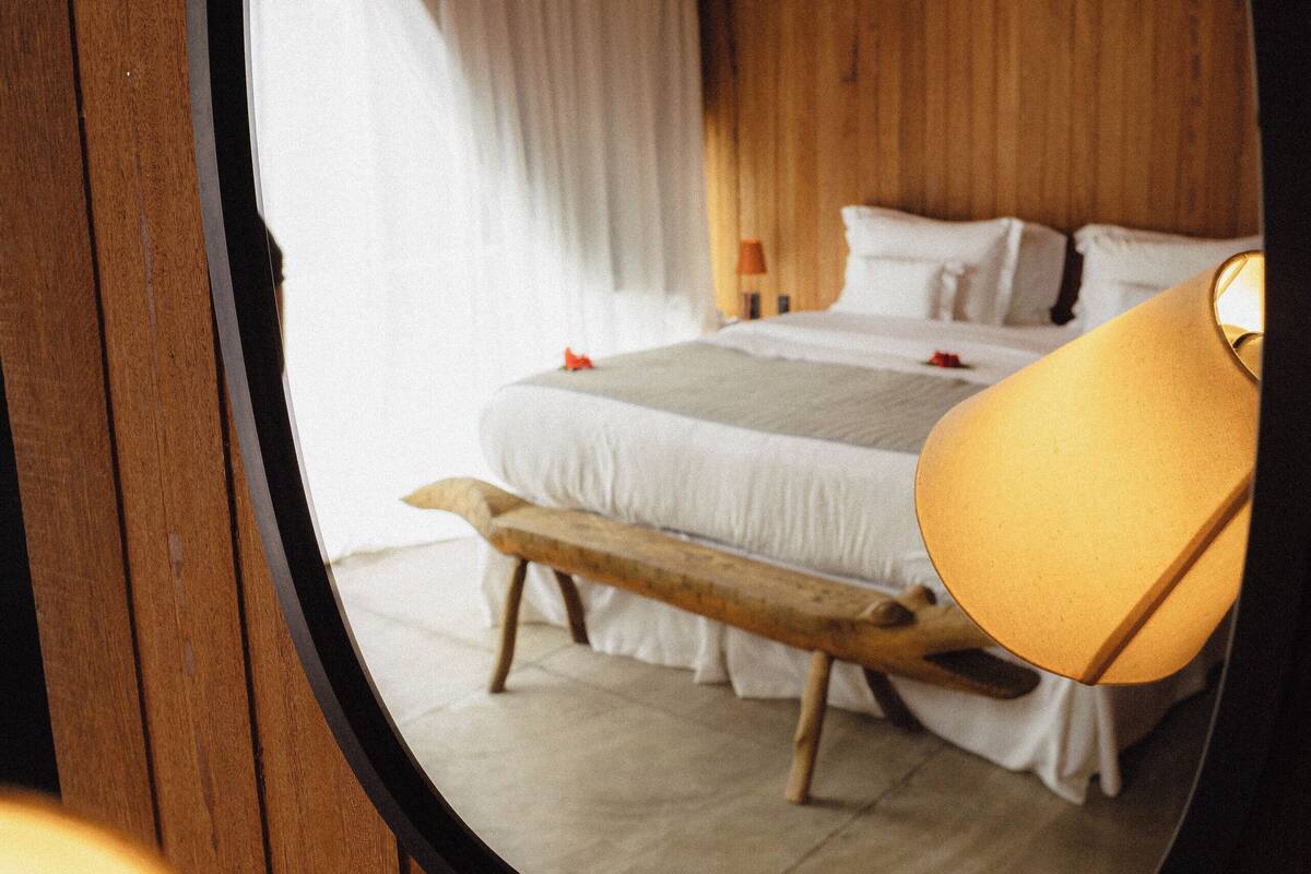 A bedroom with a wooden ceiling, a bed with white bedding, a bench, and a lamp.