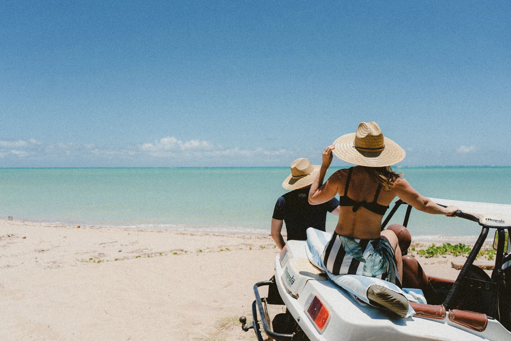 Two people on a beach, one in a bikini and the other in a t-shirt, both wearing hats, sitting on a white vehicle with a red light.
