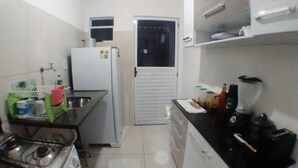 A compact kitchen with a gas stove, a white refrigerator, and a sink. There are cabinets and a countertop with various items. - Casa póxima ao Aeroporto e UFAL (Maceió)