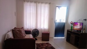A living room with a sofa, ottoman, fan, and a TV on a stand. - Casa póxima ao Aeroporto e UFAL (Maceió)