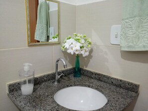 Une salle de bain avec un lavabo doté d’un plan de travail en granit, un miroir avec un cadre doré, un vase vert contenant des fleurs blanches et un distributeur de savon.