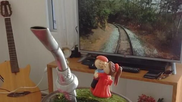 A person holding a traditional yerba mate cup with a decorative figure, a guitar, and a television in the background. - Casa póxima ao Aeroporto e UFAL (Maceió)
