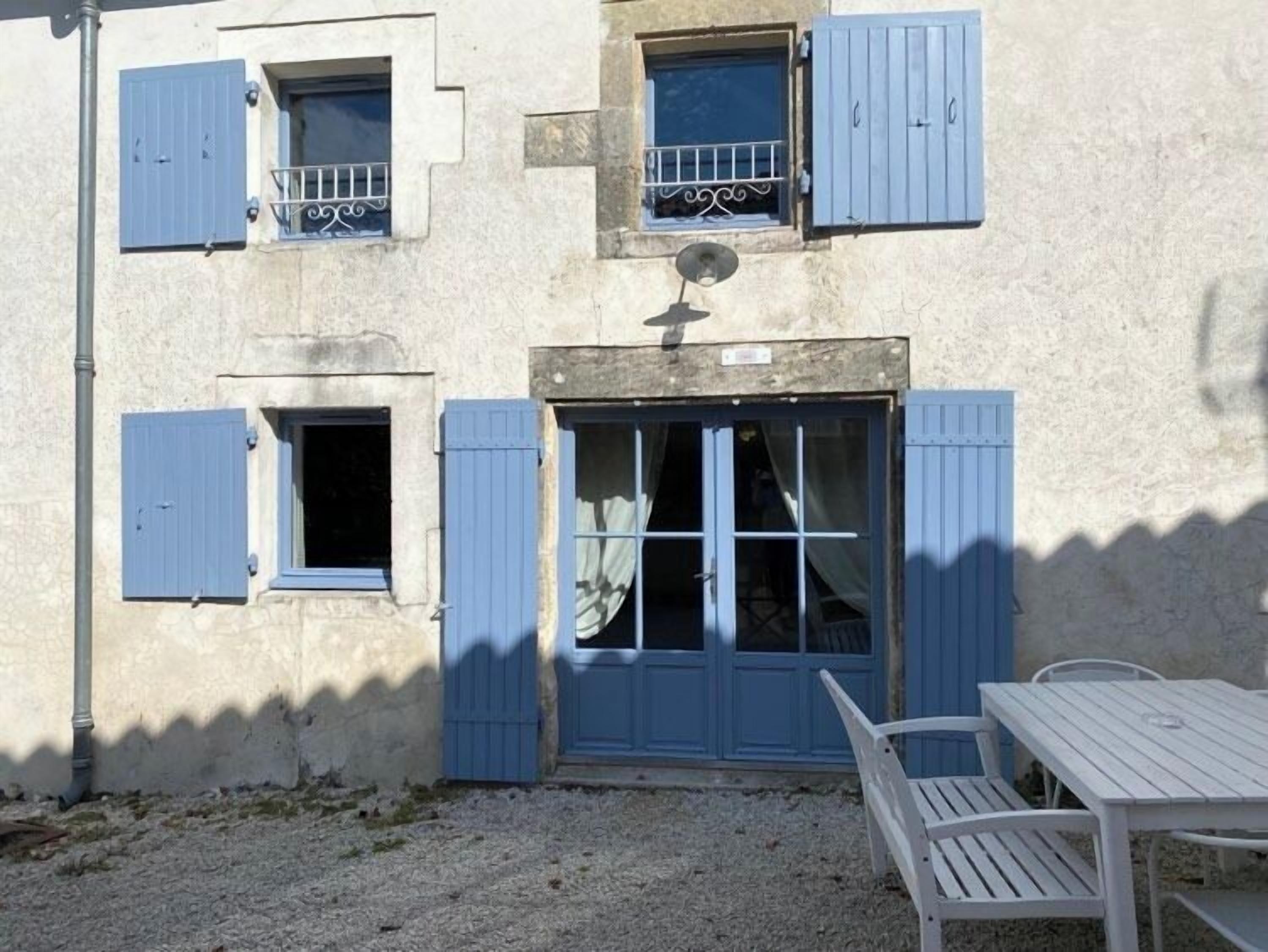 A building with blue shutters, a door, and a small window with a metal railing. There is a white table and a bench outside.