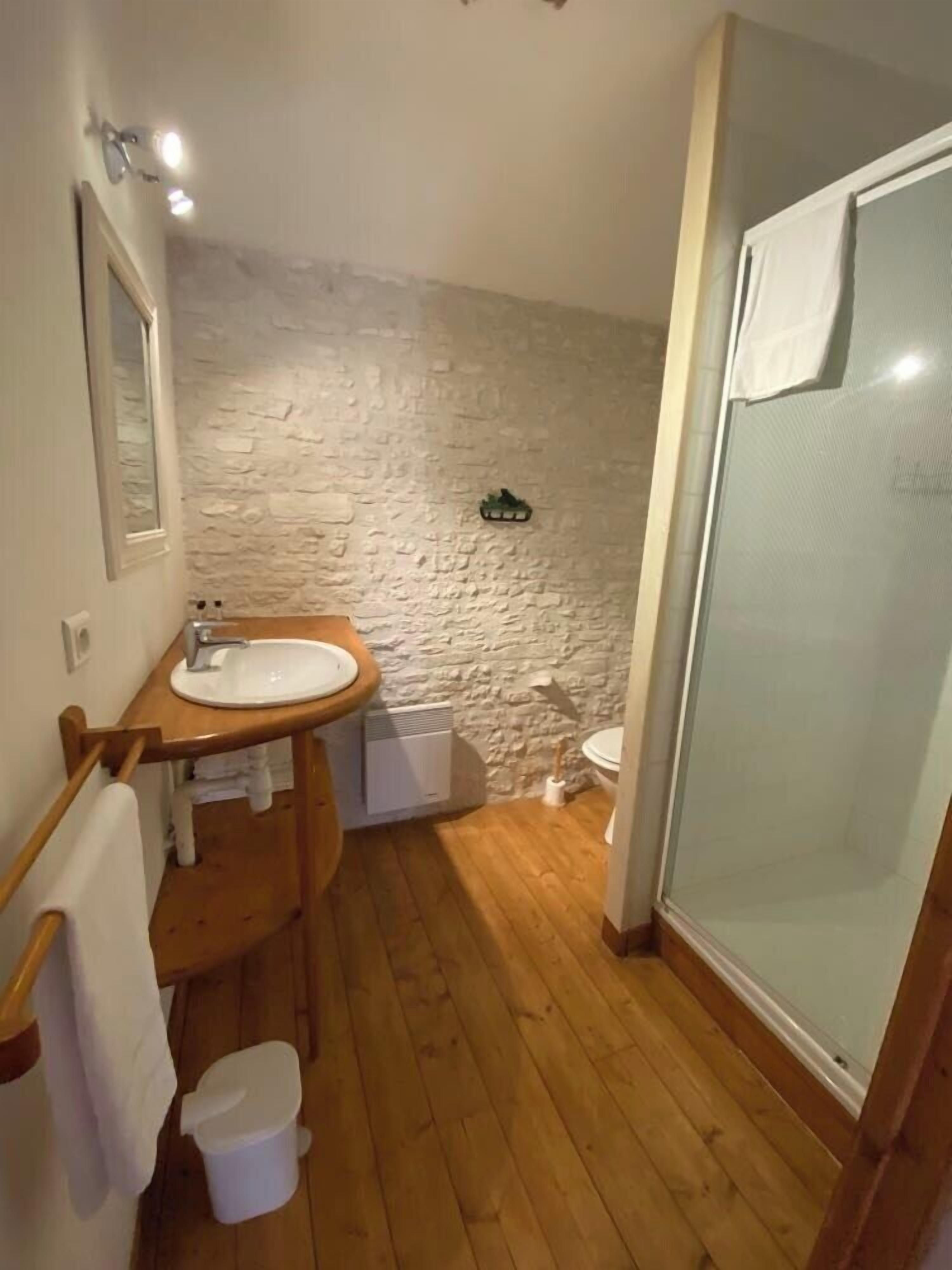 Une salle de bain avec un plancher en bois, un mur en pierre, un lavabo, des toilettes et une cabine de douche en verre.