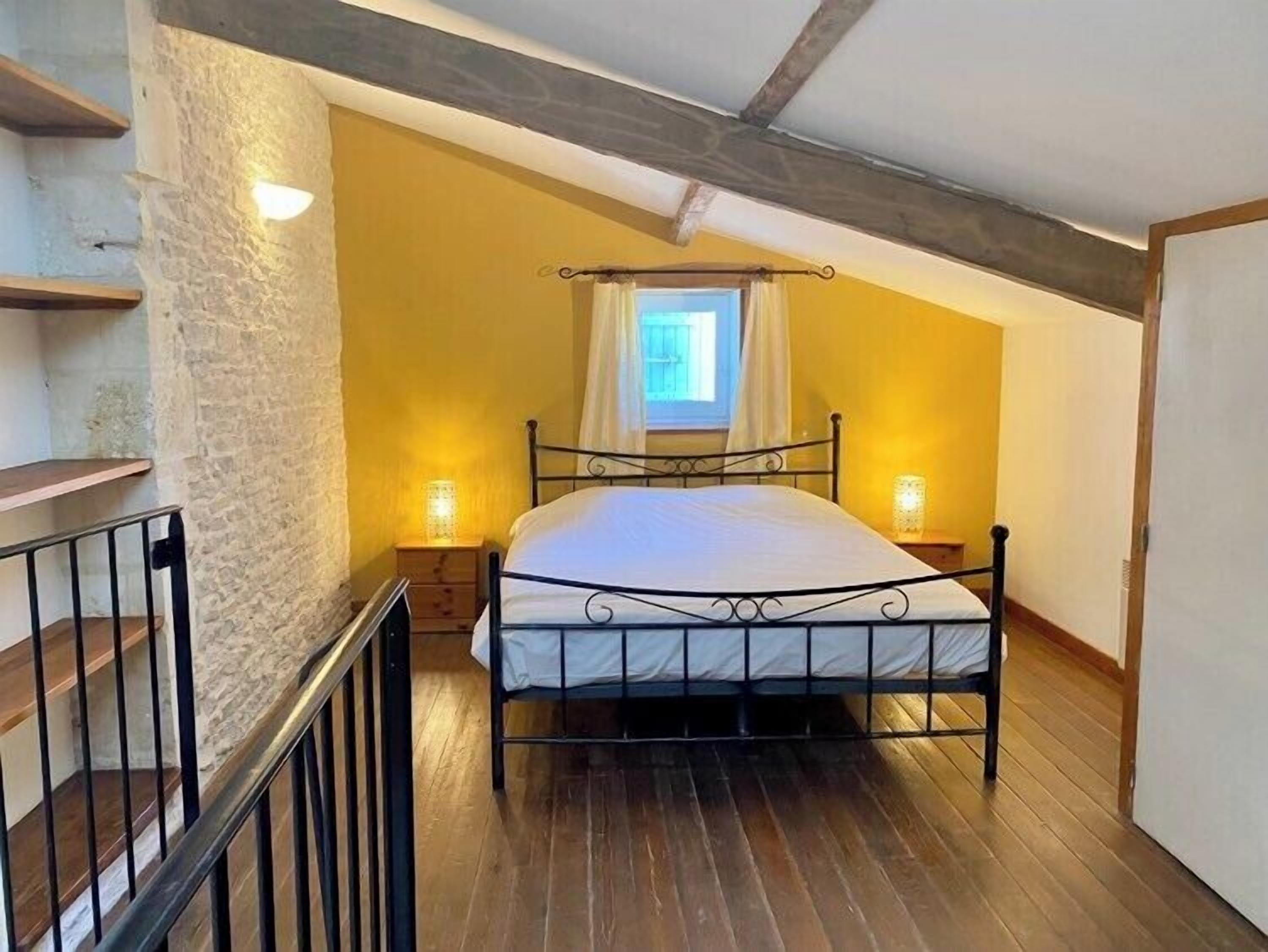 A bedroom with a sloped ceiling, a bed with a metal frame, two bedside lamps, and a window with curtains.
