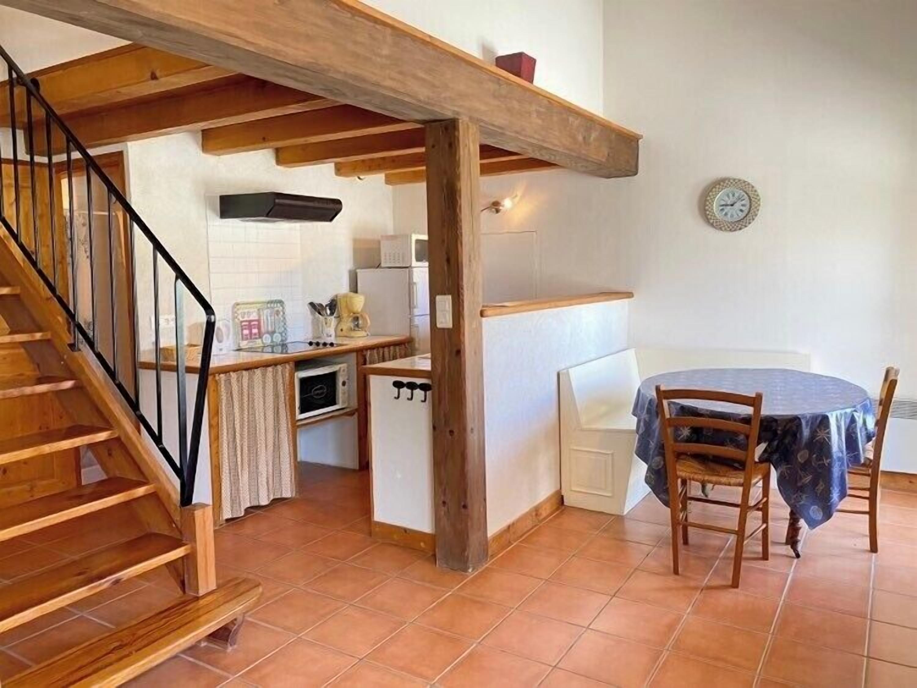 Een kleine keuken met houten balken, een eethoek met een ronde tafel en stoelen, en een trap met houten leuningen.