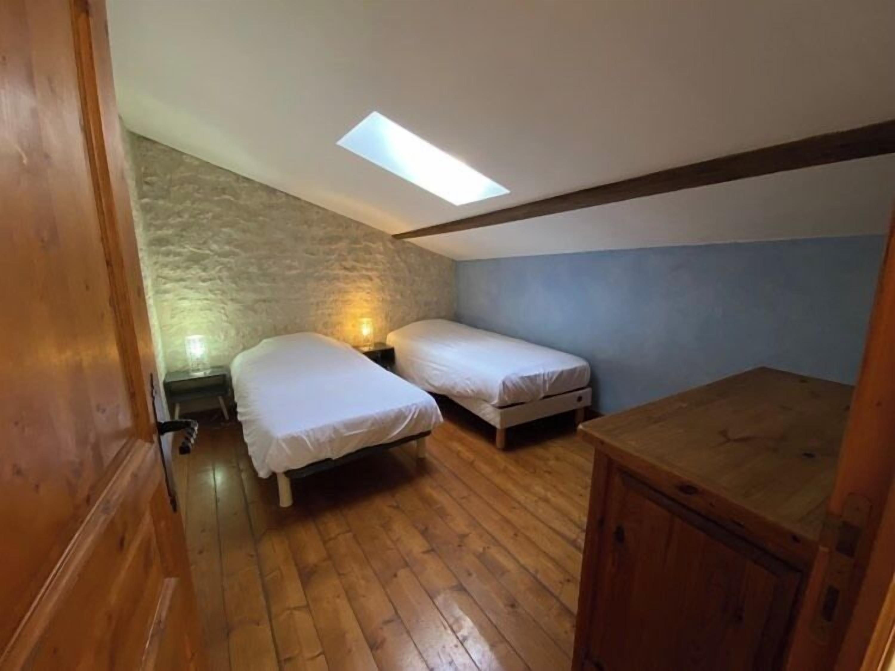 A small, cozy room with two single beds, a wooden floor, a sloped ceiling with a skylight, and a stone wall.