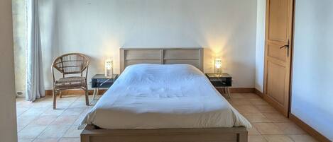 A bedroom with a wooden bed, two bedside tables with lamps, a wicker chair, and a wooden door.