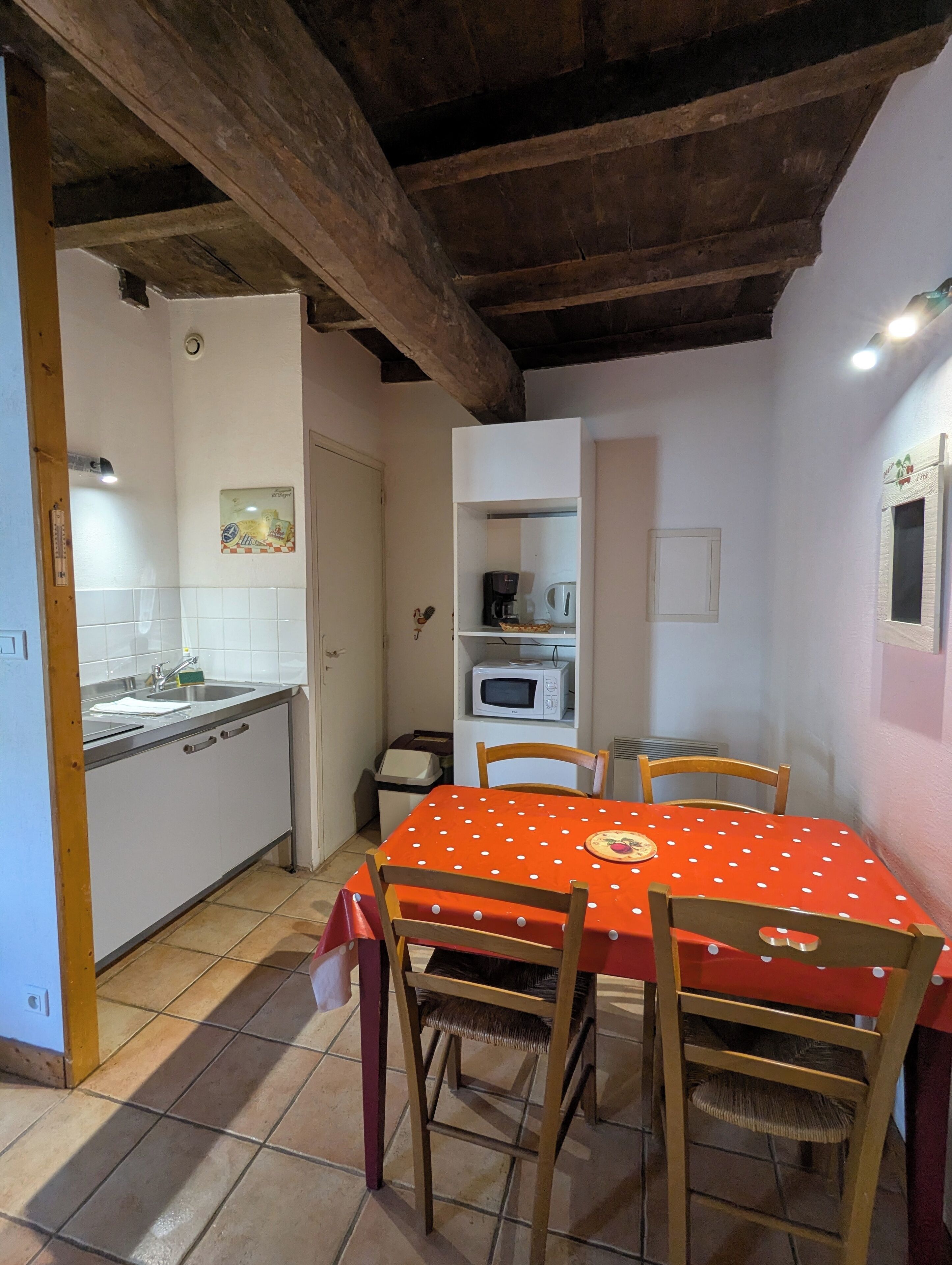 A kitchen with a white microwave, a coffee maker, and a sink. There is a dining table with a red tablecloth and four chairs. The room has wooden beams on the ceiling and white walls.