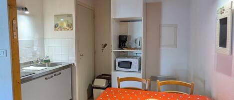 Una cocina con microondas blanco, cafetera y lavamanos. Hay una mesa de comedor con mantel rojo y cuatro sillas. La habitación tiene vigas de madera en el techo y paredes blancas.