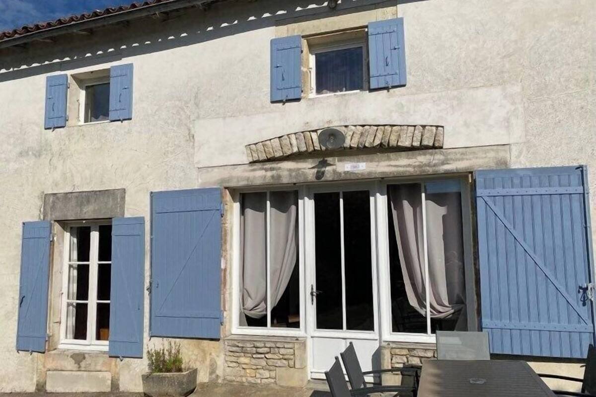 Ein Gebäude mit zahlreichen Fenstern, einige davon mit Fensterläden, und einem steinernen Rundbogen über dem Haupteingang. Vor der Tür stehen ein Tisch und Stühle.