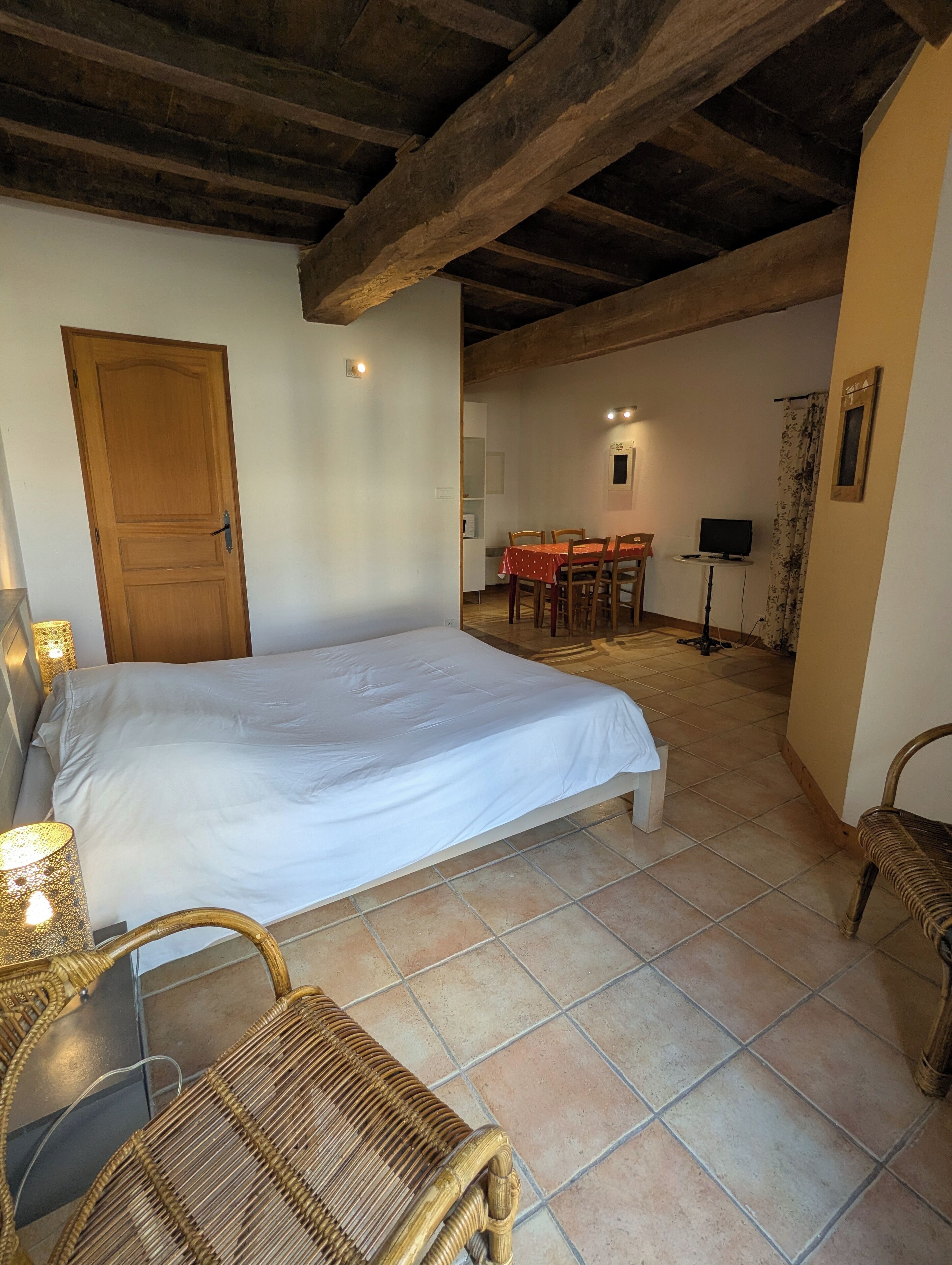 Une chambre avec un grand lit, des poutres apparentes en bois au plafond, une table à manger avec des chaises et un téléviseur dans l’espace repas.