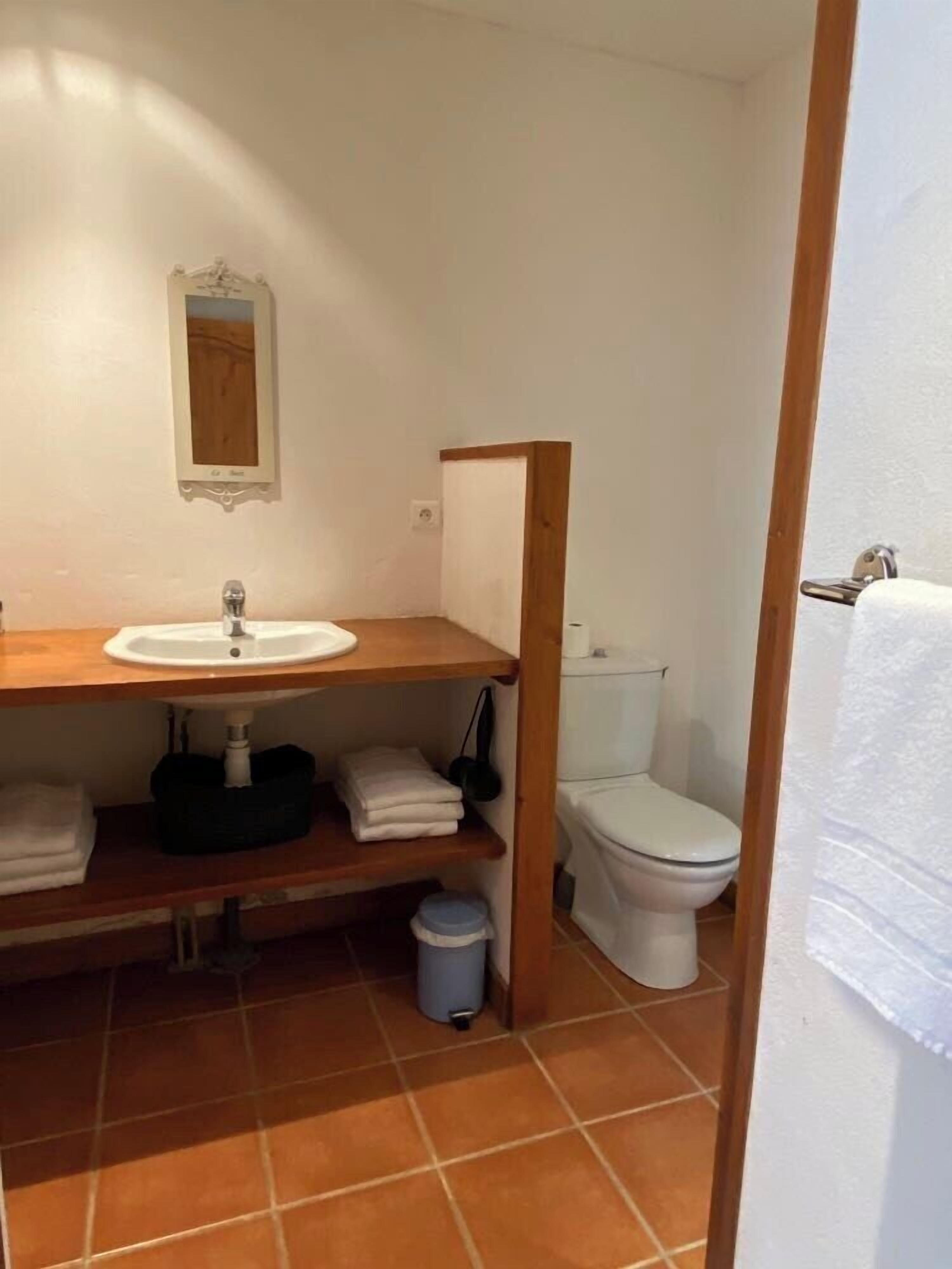A small bathroom with a wooden vanity, a white sink, a toilet, and a small trash bin.
