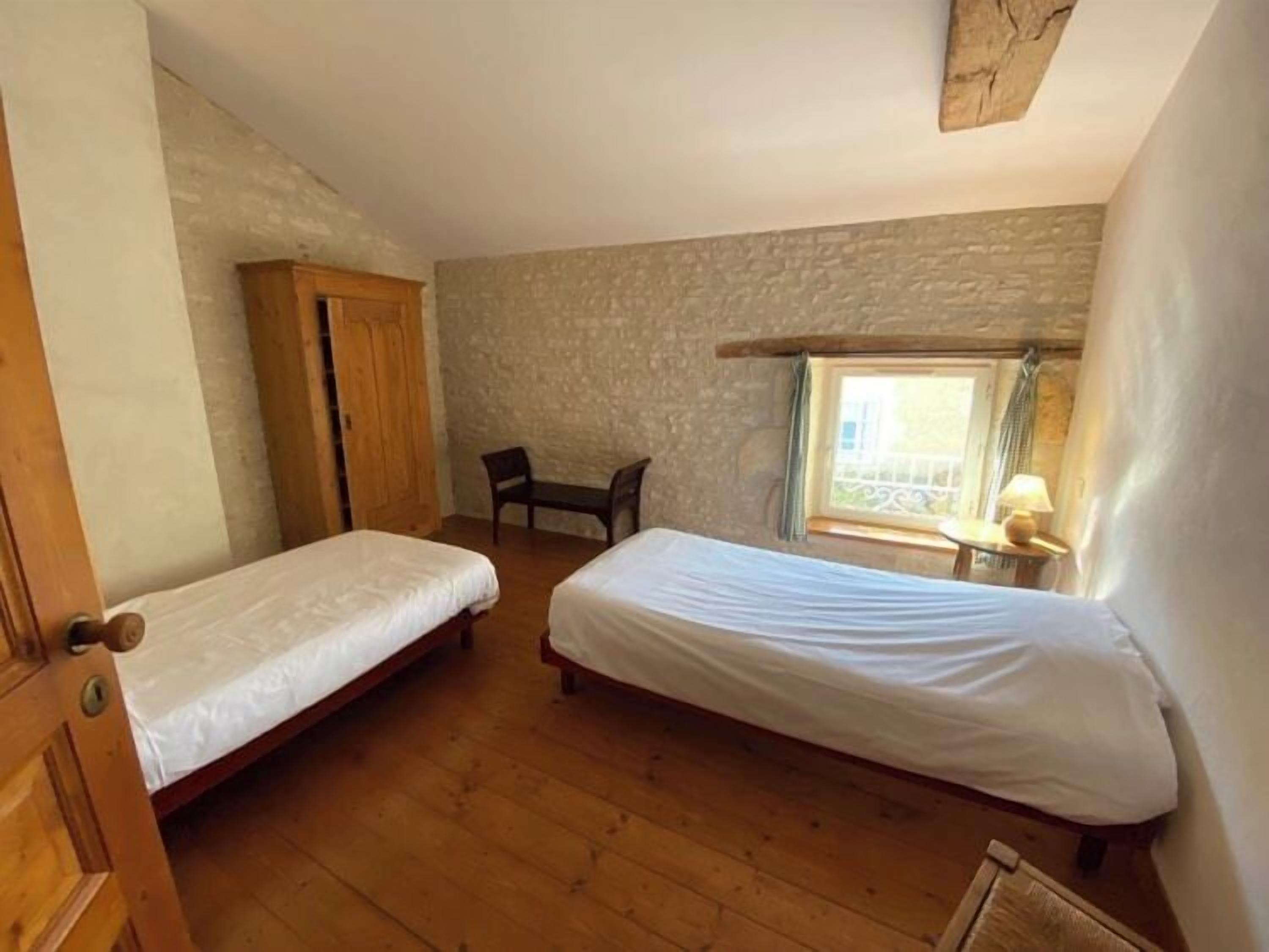 A room with two single beds, a wooden wardrobe, a small table with a lamp, and a window with a view of the outside.