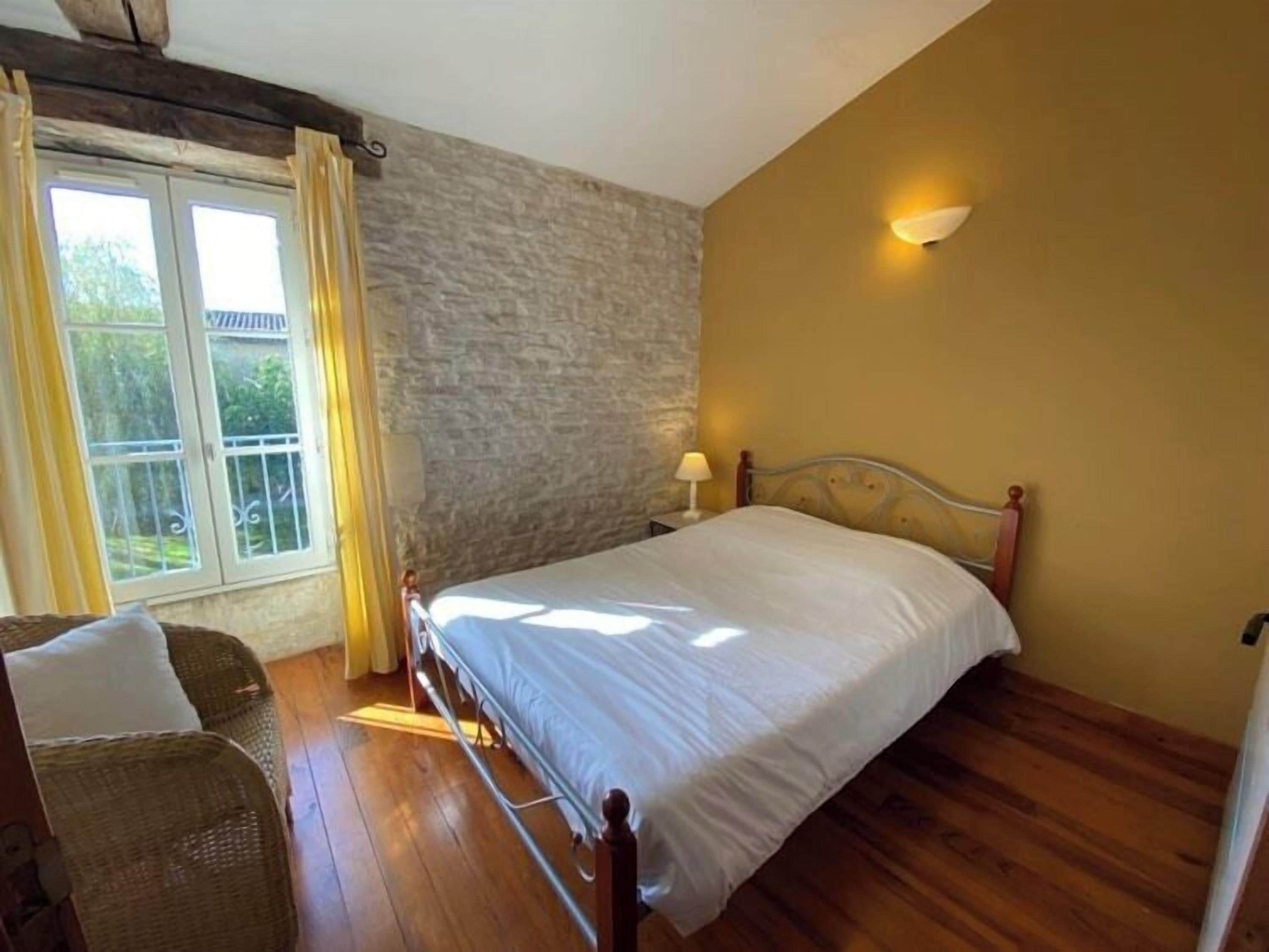Un dormitorio con cama, mesita de noche, lámpara, sillón de mimbre y ventana con cortinas.