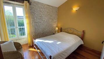 Un dormitorio con cama, mesita de noche, lámpara, sillón de mimbre y ventana con cortinas.