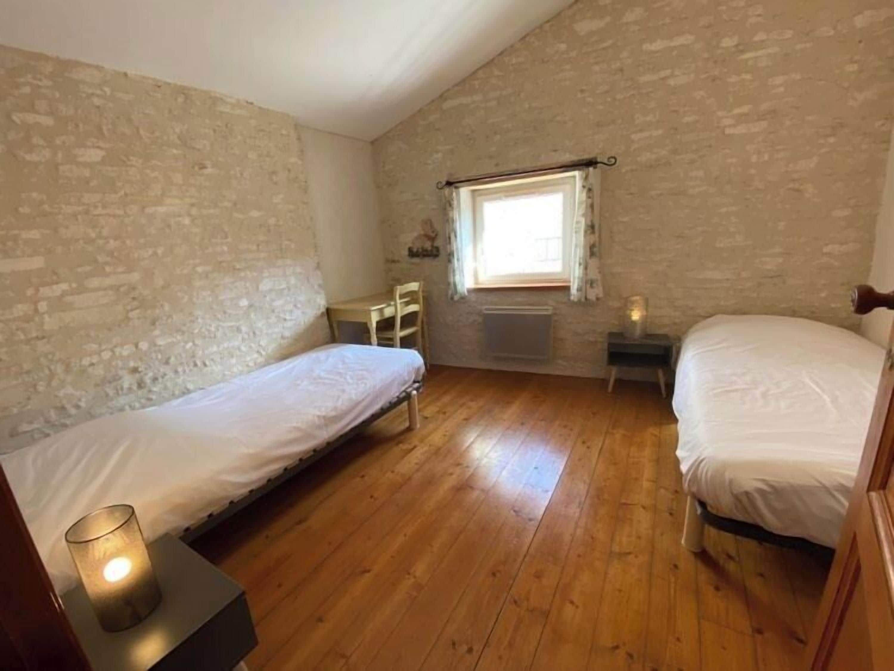 Una habitación pequeña y rústica con dos camas individuales, piso de madera, una ventana con cortinas y una mesita con una lámpara.