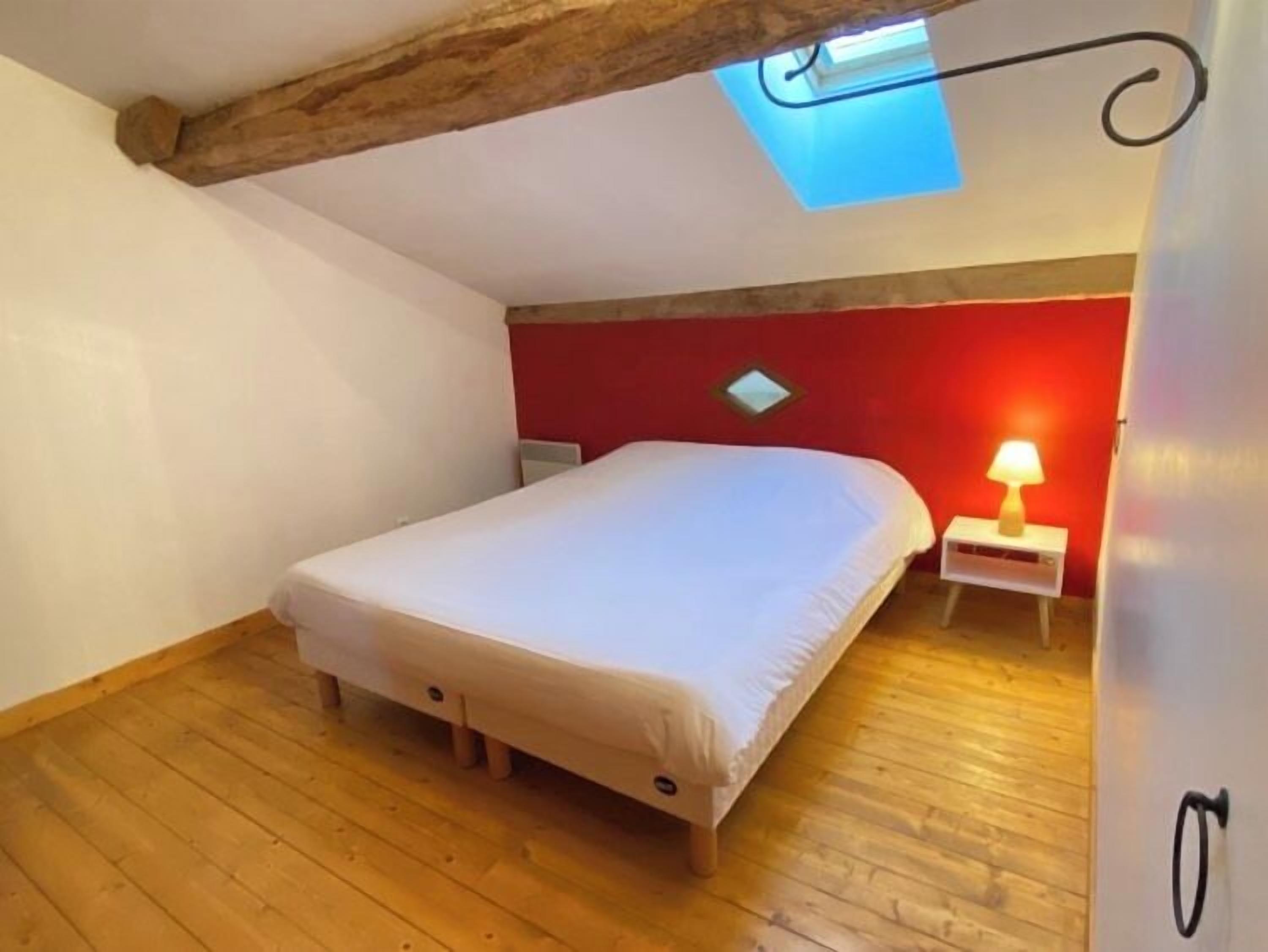 Une chambre à coucher comprenant un lit, une table de chevet, une lampe, un mur d’accent rouge, un plancher en bois et un puits de lumière.