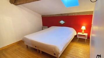 Une chambre à coucher comprenant un lit, une table de chevet, une lampe, un mur d’accent rouge, un plancher en bois et un puits de lumière.