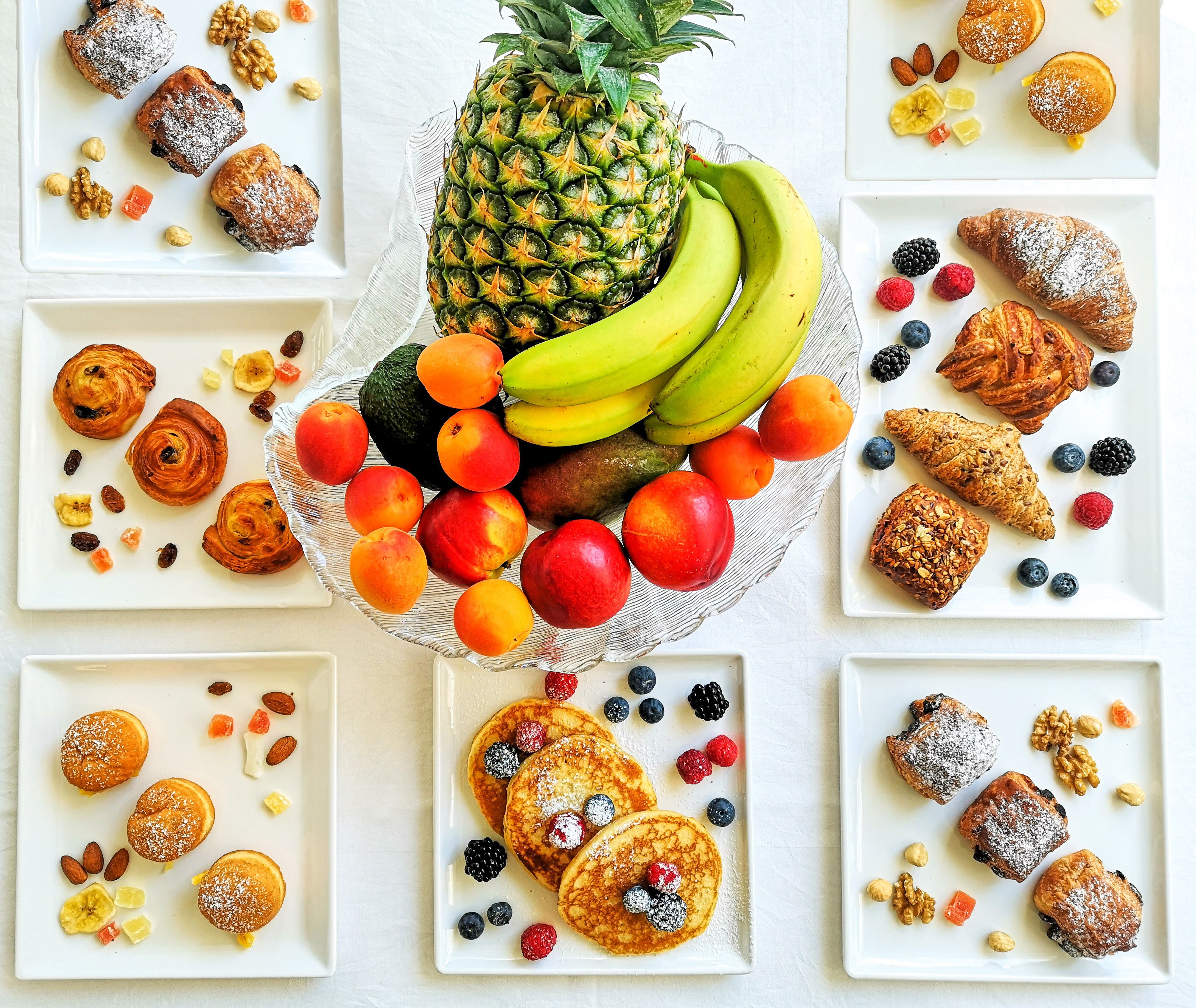 Una bandeja de frutas con plátanos, piñas y diversas bayas, rodeada de platos con pasteles y muffins.