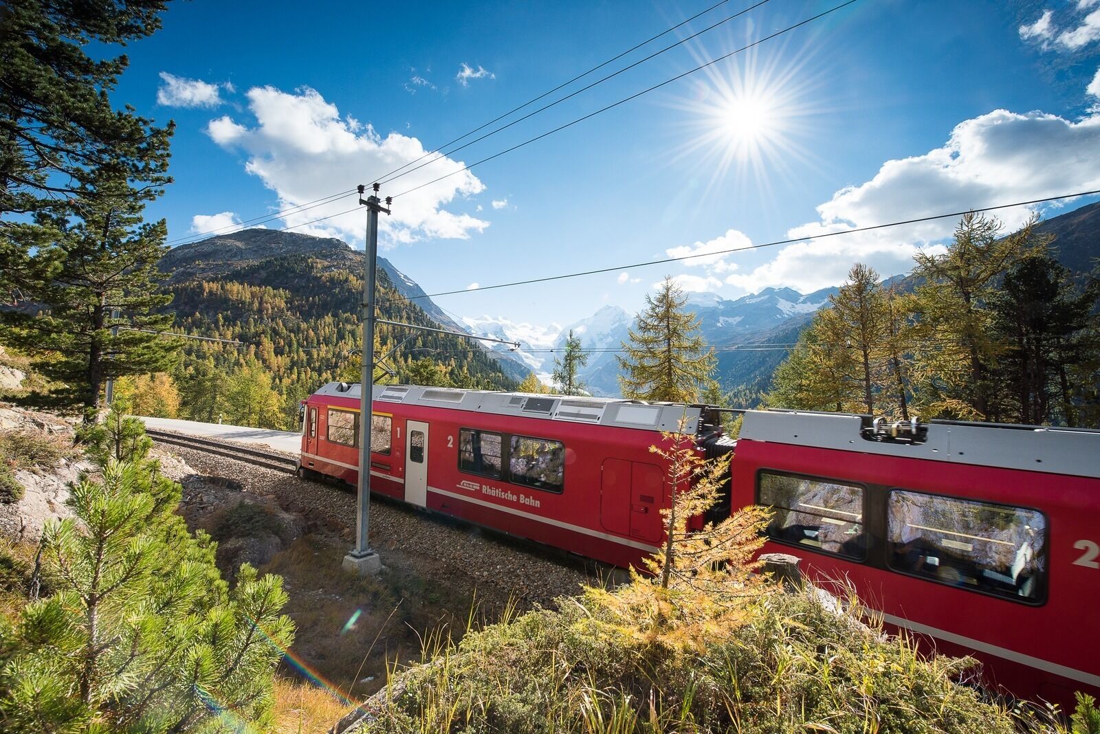 A red train with "Rheinfahrt Bahn" written on the side, traveling through a mountainous area with pine trees and a clear sky.