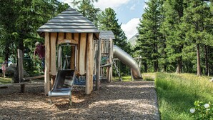 A playground with a slide and climbing structures, surrounded by trees and a gravel path. - Hotel Morteratsch (Pontresina)