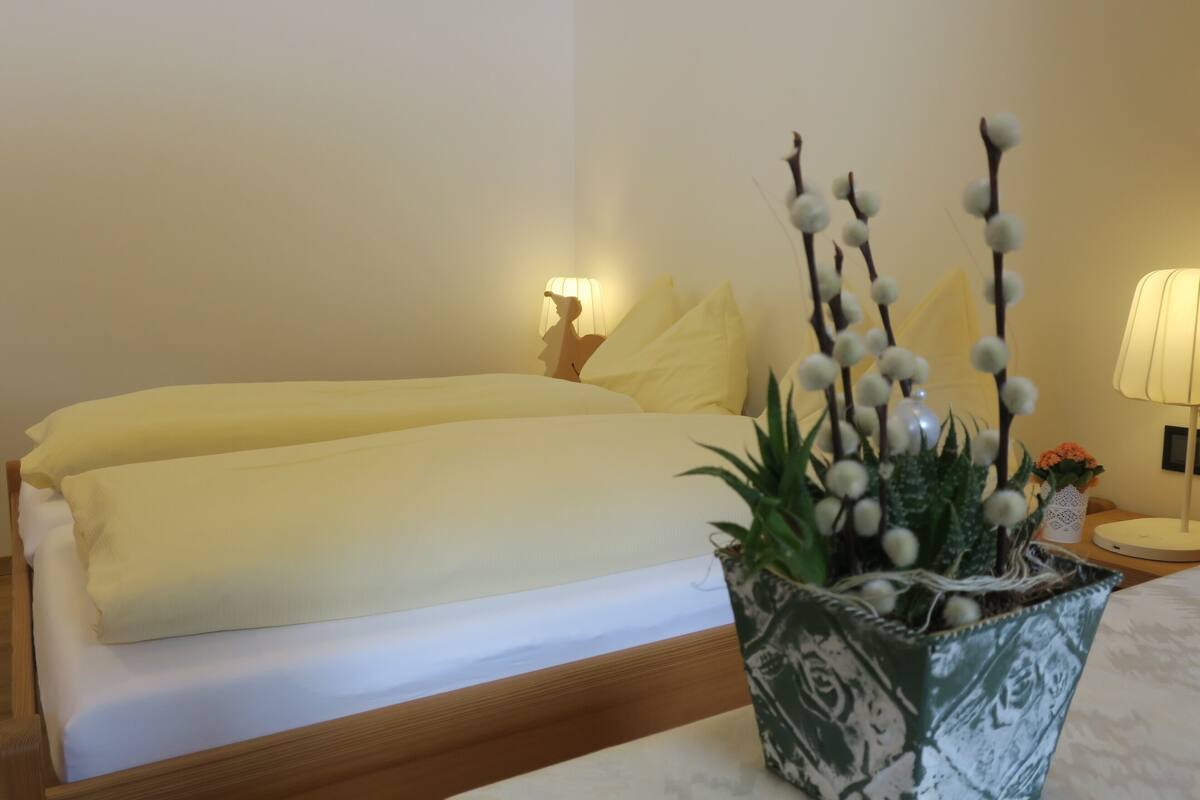 A neatly made bed with a yellow blanket, a bedside lamp, and a potted plant with white flowers.