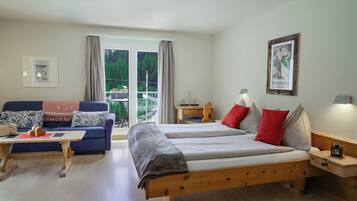 Una habitación con dos camas, un sofá, una mesa y una silla. Hay una ventana con cortinas y vistas a zonas verdes.