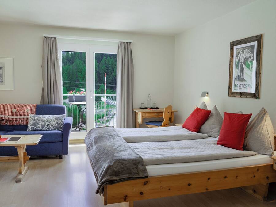 Una habitación con dos camas, un sofá, una mesa y una silla. Hay una ventana con cortinas y vistas a zonas verdes.