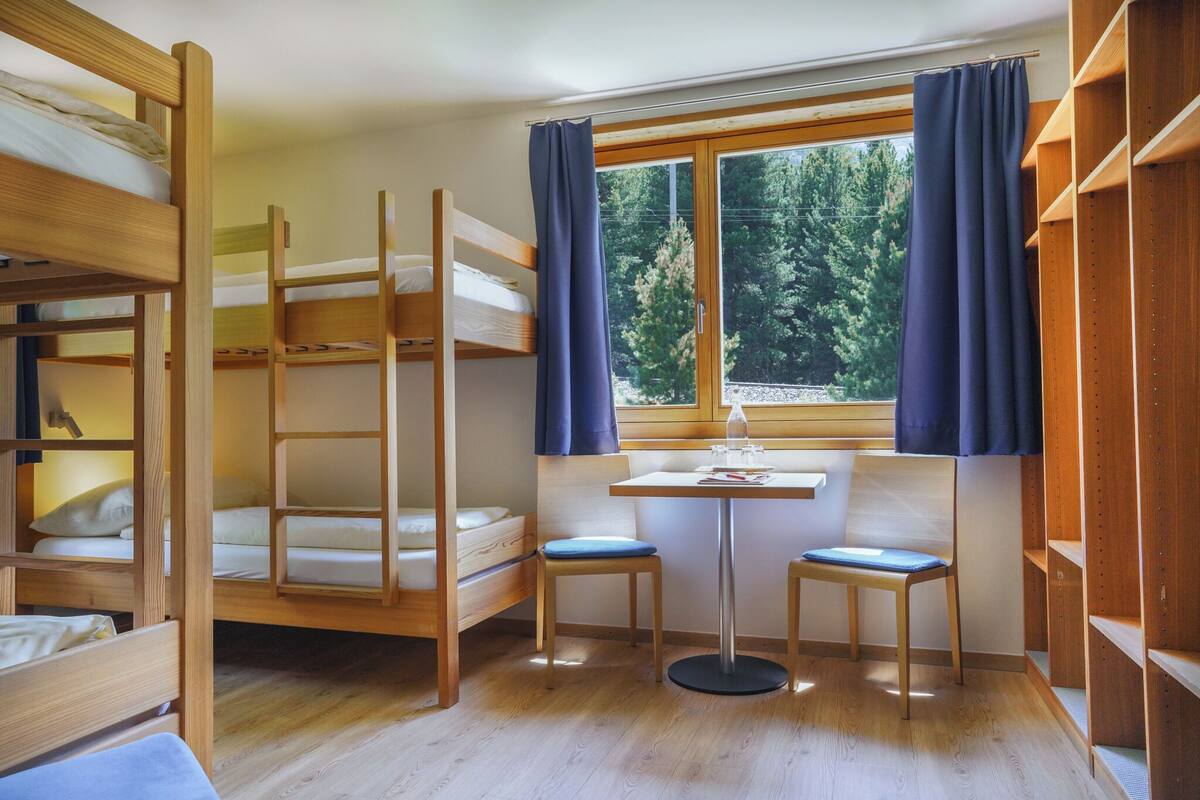 A dormitory room with bunk beds, a small table with chairs, and a window with curtains.