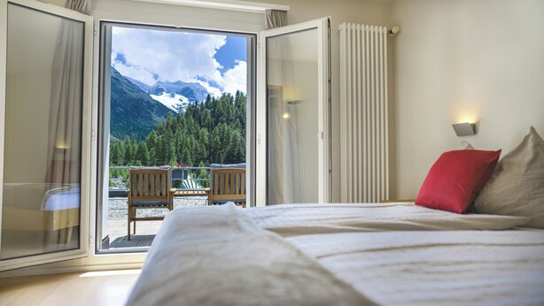 A bedroom with a bed, a red pillow, and a view of a mountain landscape through a glass door. - Hotel Morteratsch (Pontresina)