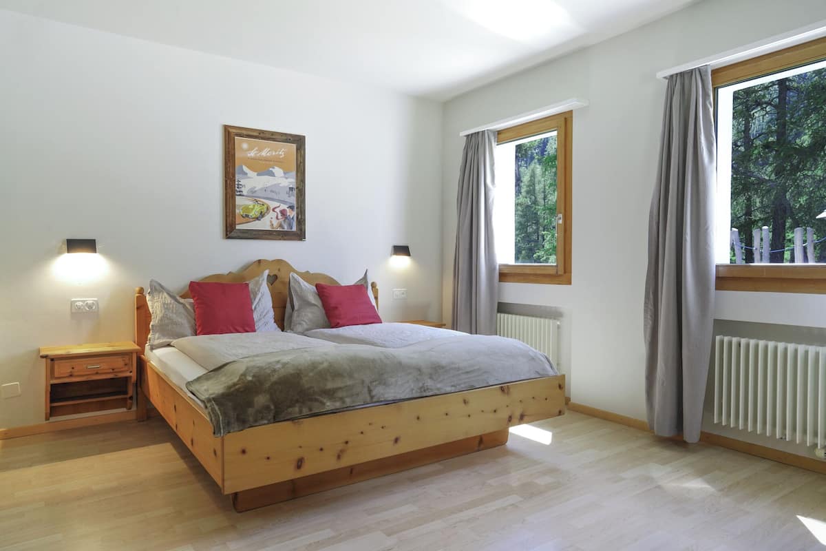 A bedroom with a wooden bed, two bedside tables, a nightstand, a window with curtains, and a wall-mounted picture.