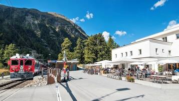 A train station with a red train, outdoor seating, and a mountain backdrop.