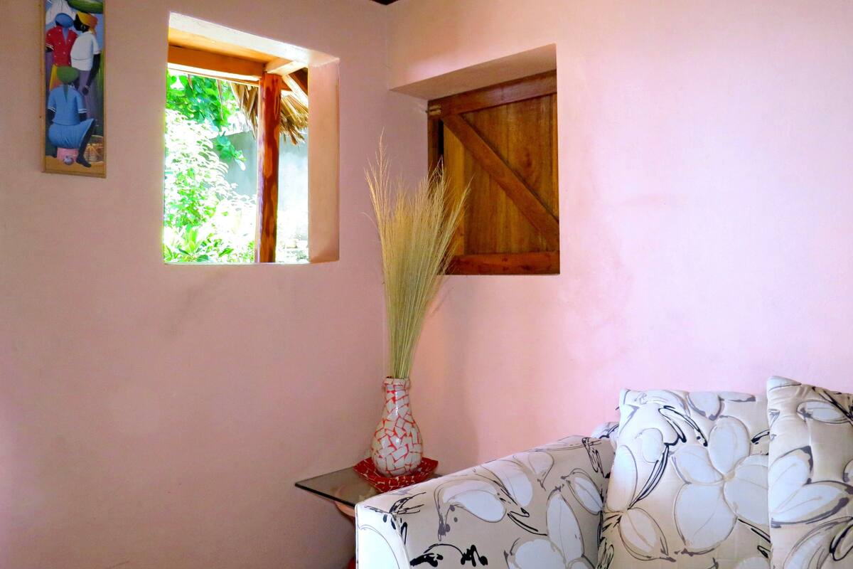 Une pièce avec un mur rose, une petite fenêtre donnant sur un espace verdoyant, une porte en bois et un canapé blanc à motifs.