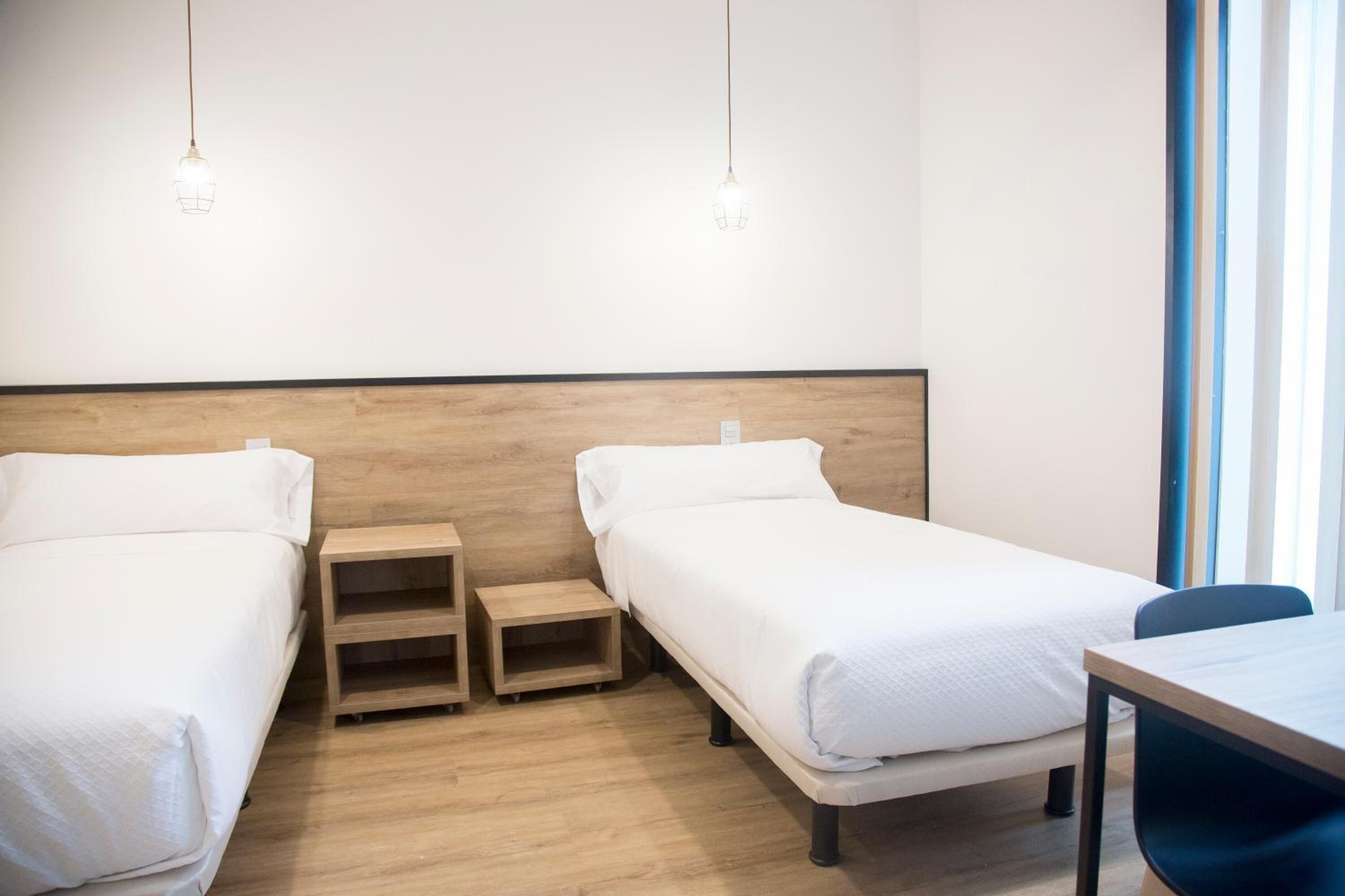 A hotel room with two single beds, a wooden nightstand, a small table, and a window with blinds.
