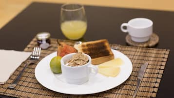 Desayuno con tostadas, cereal, fruta, queso, un vaso de jugo de naranja y una taza sobre una mesa con posavasos.