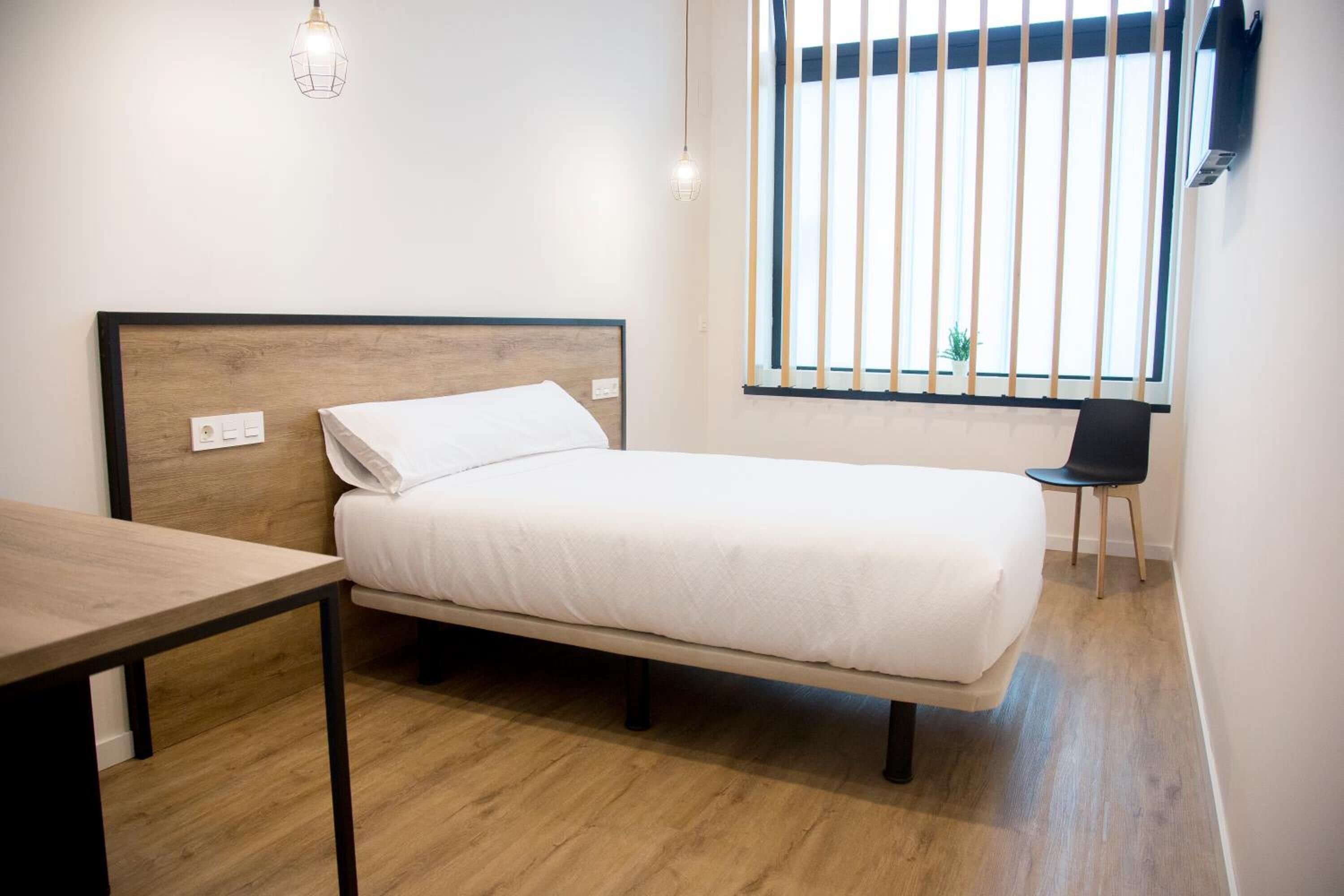 Una cama individual con cabecera de madera, una mesita de noche, una silla y una ventana con persianas verticales.