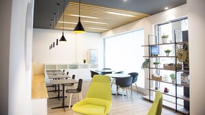 A modern office space with a dining area, multiple tables, chairs, and a bookshelf with plants.
